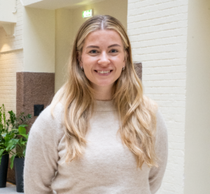 En person står innendørs med armene bak ryggen, iført en lys beige genser og mørke bukser med belte. Bakgrunnen viser en lys murvegg og grønne planter.