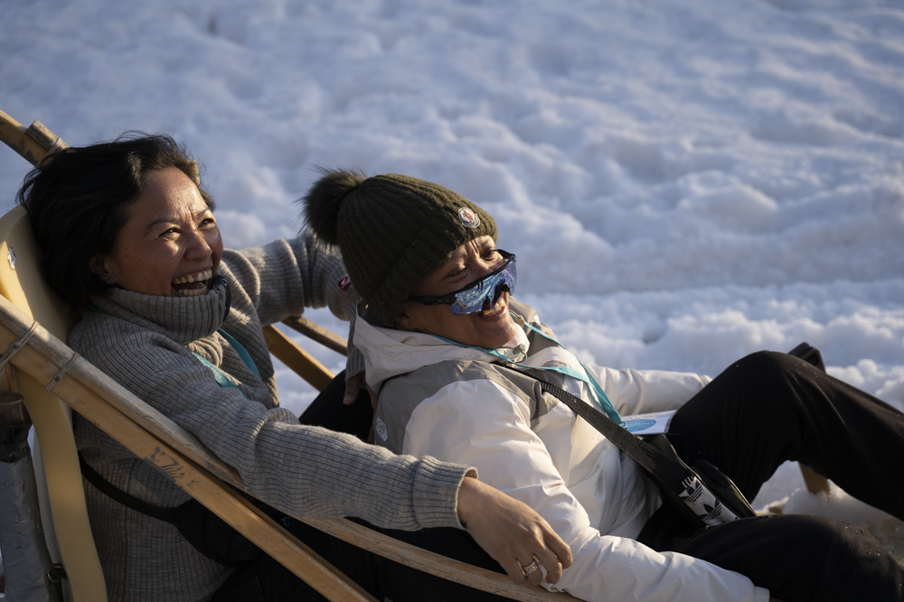 To kvinner sitter sammen i en treslede, smilende og leende, med snø på bakken rundt seg.