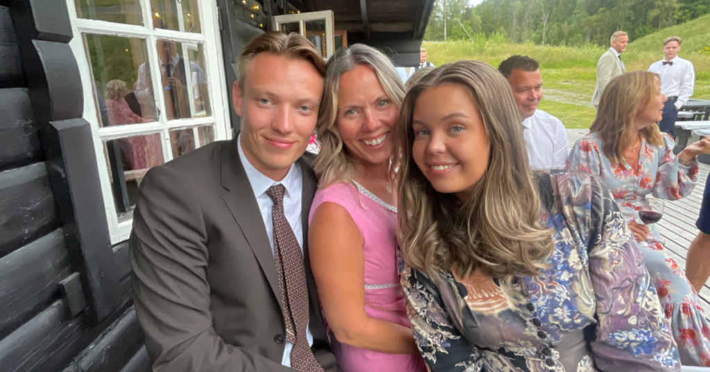 Tre formelt kledde personer sitter tett sammen og smiler til kameraet utenfor en trebygning, med andre og grøntområder synlige i bakgrunnen.