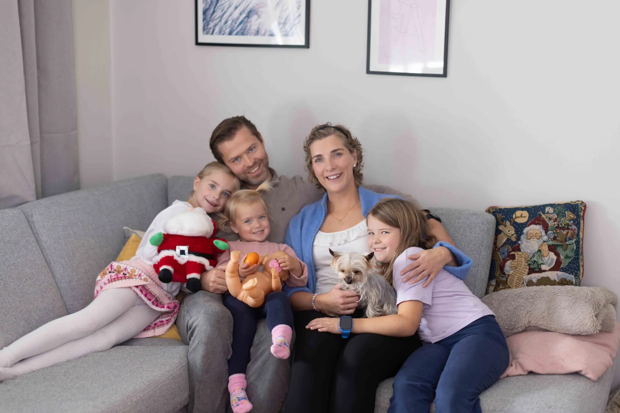 En familie på fem, to voksne, tre barn og en liten hund, sitter sammen i en grå sofa og smiler til kameraet. To barn holder i kosedyr.