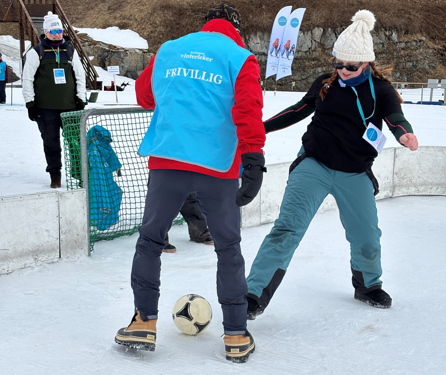 To voksne spiller fotball på snø, den ene iført blå vest med påskriften "FRIVILLIG". Et lite barn og to personer står i bakgrunnen.