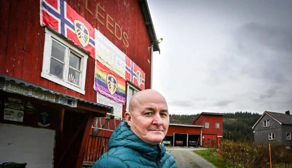 En mann i blågrønn jakke står foran en rød bygning dekorert med Leeds United og norske flagg, samt et regnbueflagg. Andre hus og trær er synlige i bakgrunnen.