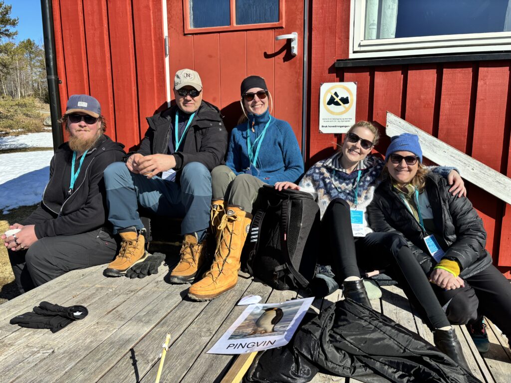 Fem personer sitter på et tredekke utenfor en rød bygning, iført kalde klær og støvler, med snøen synlig i bakgrunnen. Et skilt og et papir med påskriften "PINGVIN" er til stede.