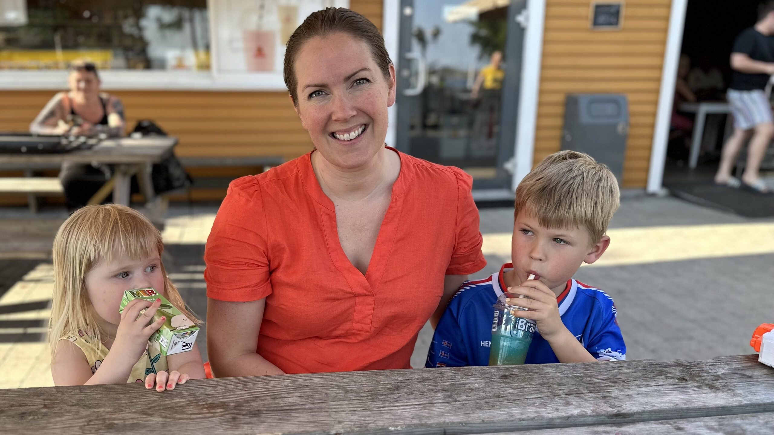 En kvinne i oransje skjorte sitter ved et utendørs trebord sammen med en ung jente og en gutt, som begge drikker av juicebokser. En gul bygning ligger i bakgrunnen.