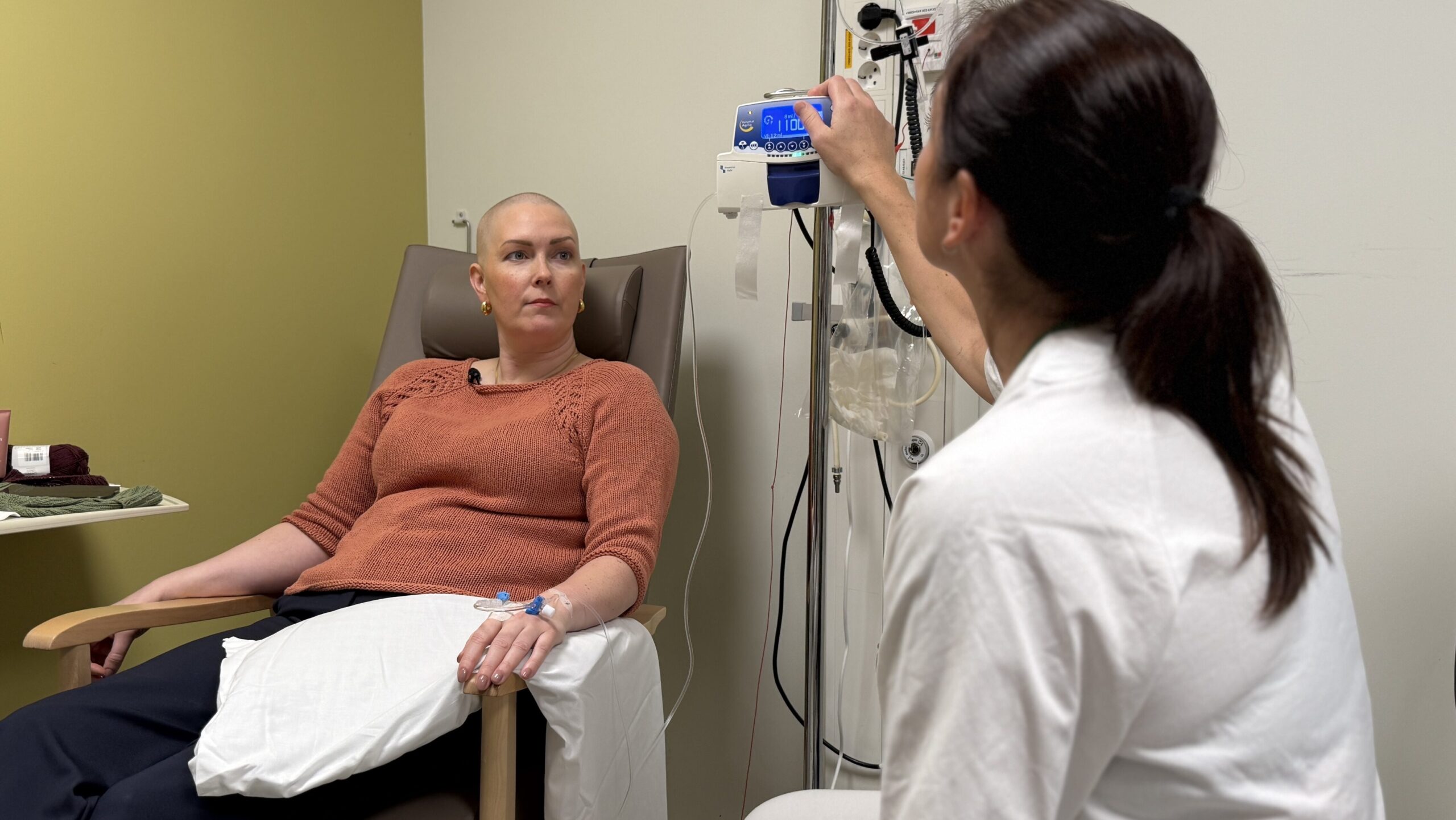 En pasient får intravenøs behandling mens hun sitter ned og en helsearbeider justerer et medisinsk utstyr ved siden av henne i en klinisk setting.