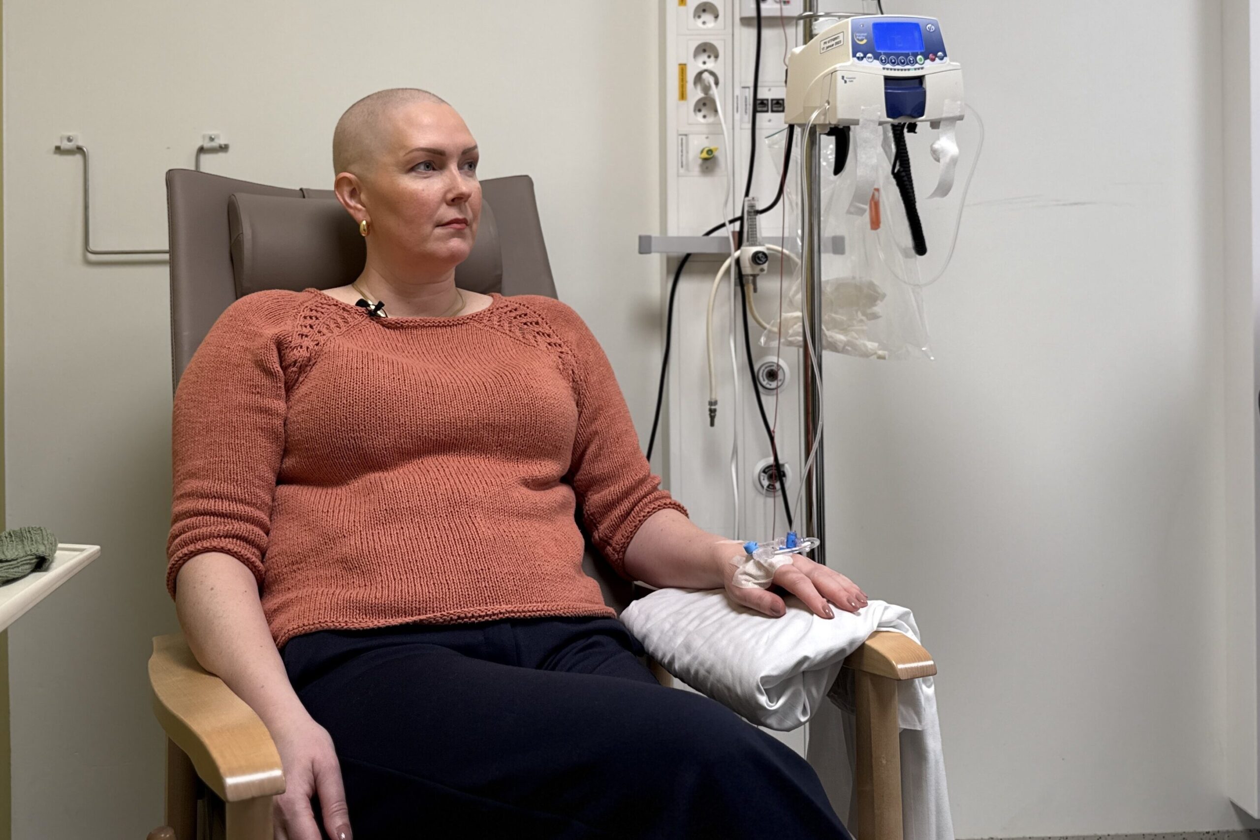 En kvinne med sitter i en sykehusstol og får intravenøs behandling, med medisinsk utstyr synlig ved siden av henne.