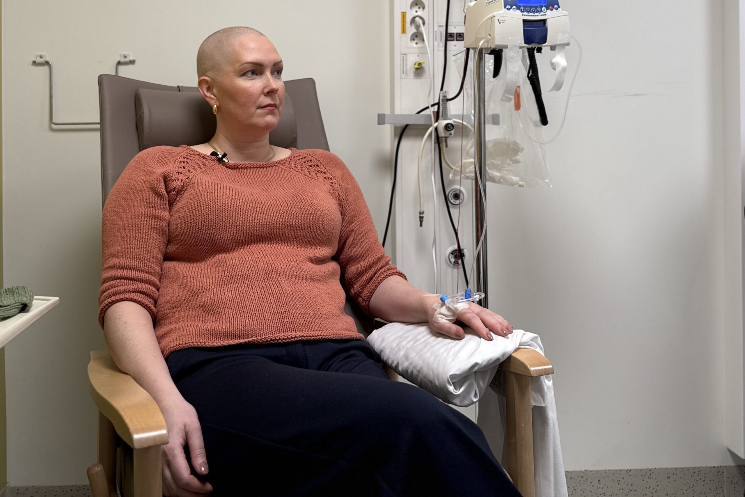 En kvinne med sitter i en sykehusstol og får intravenøs behandling, med medisinsk utstyr synlig ved siden av henne.