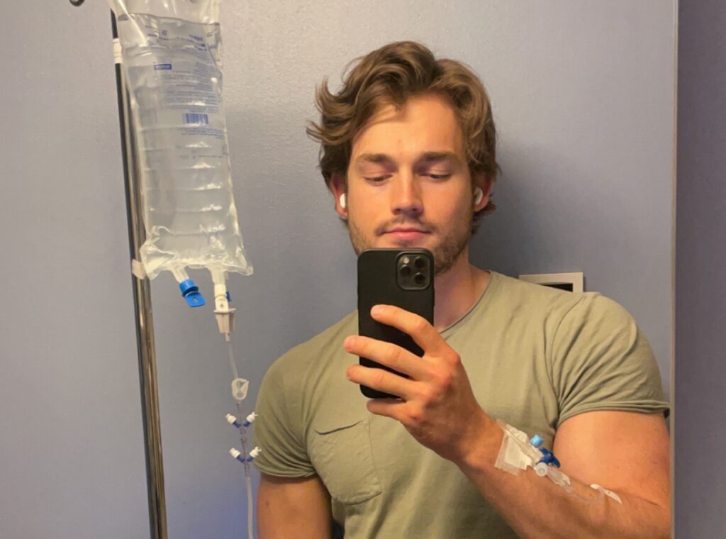 En mann står foran et speil og tar en selfie mens han er koblet til et intravenøst drypp i et medisinsk miljø.