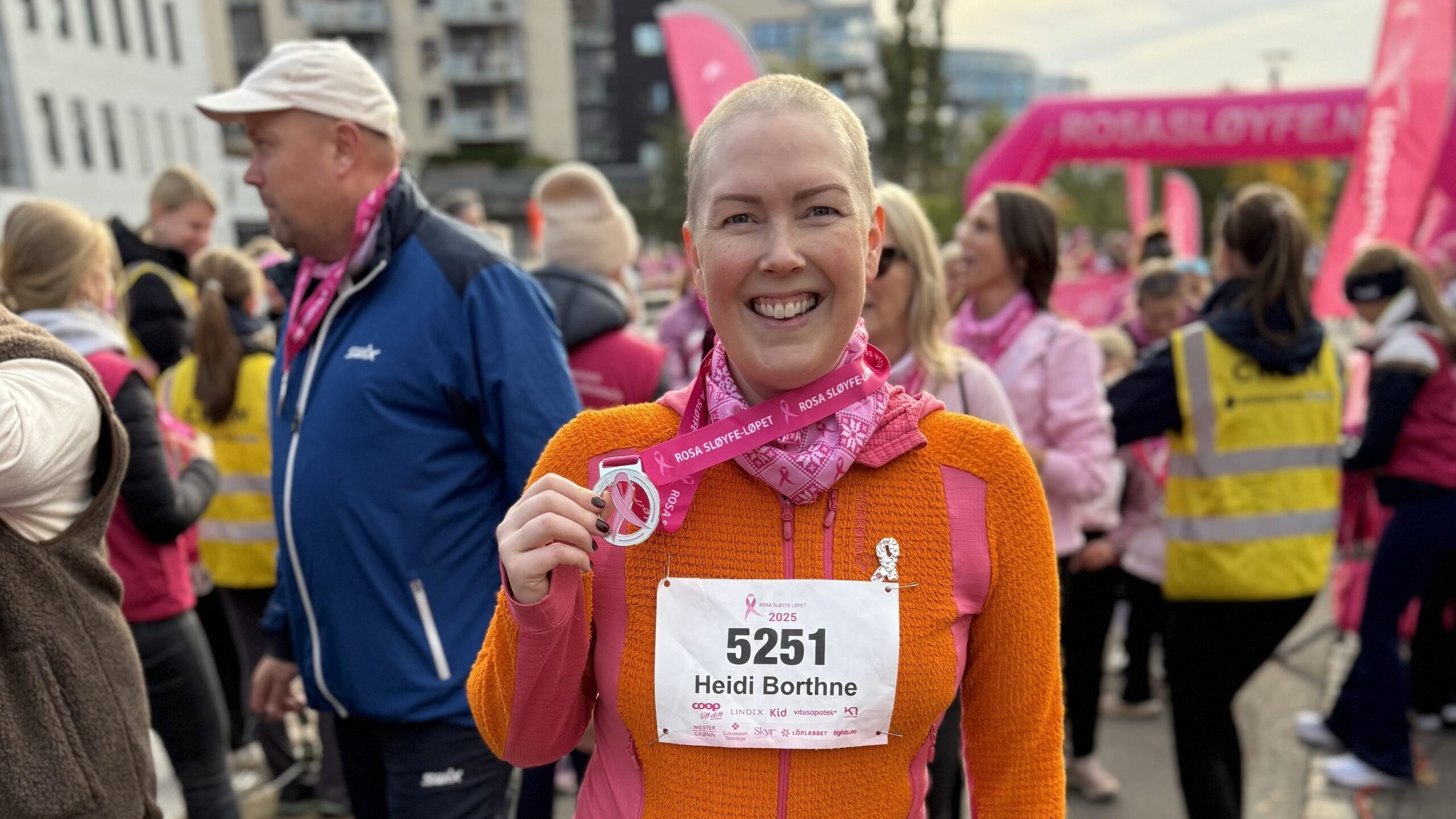 En kvinne med startnummer og medalje smiler under et Rosa sløyfe-løp for å sette fokus på brystkreft, omgitt av deltakere i rosa klær og frivillige.