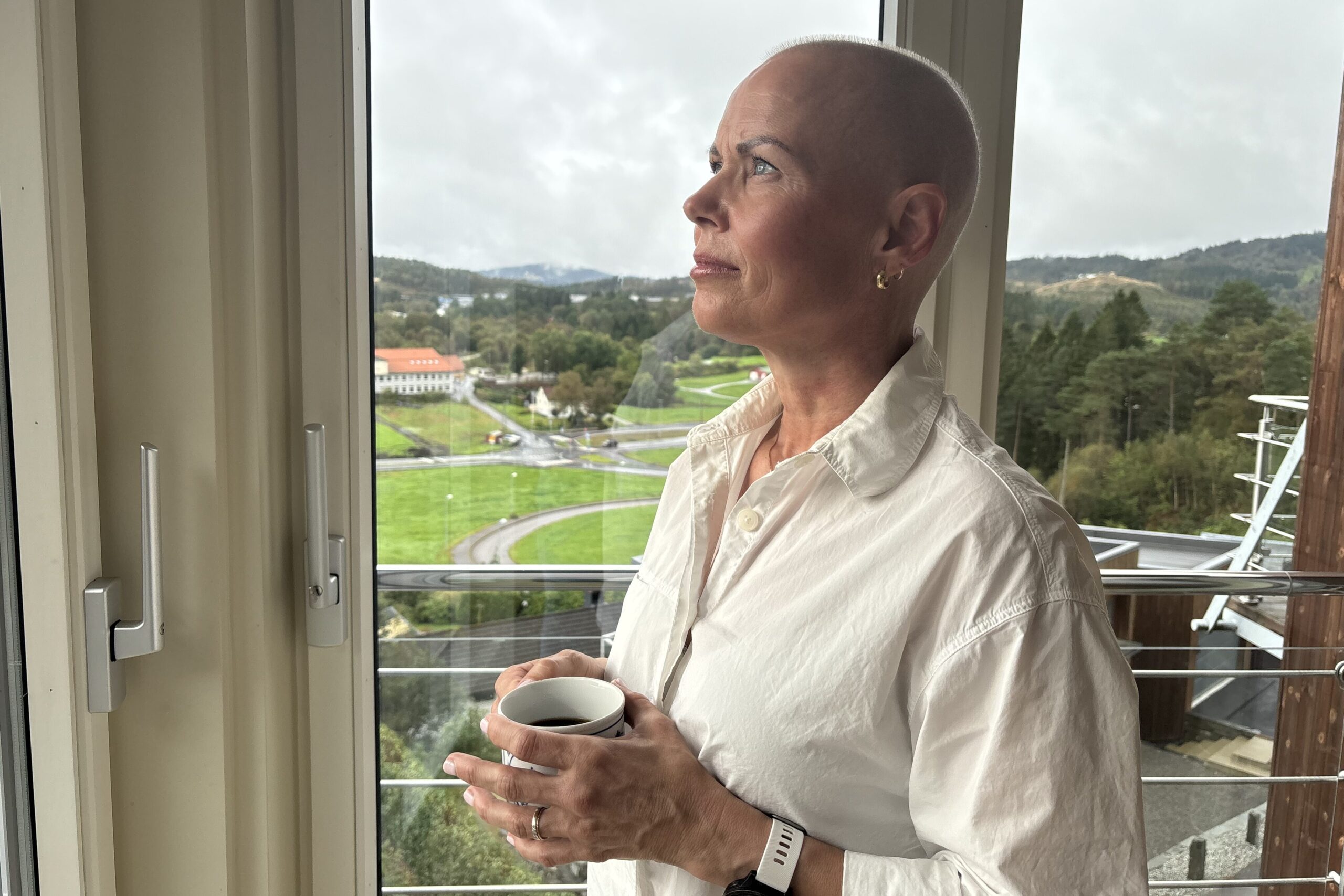 En person med barbert hode og hvit skjorte står ved et vindu med et krus i hånden og ser ut på et naturskjønt, grønt landskap.