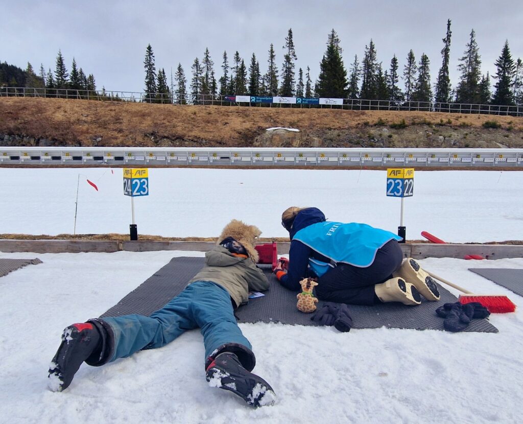 To vinterkledde personer ligger på matter i snøen på en skytebane og sikter mot skive nummer 22 og 23, med trær og tribuner i bakgrunnen.