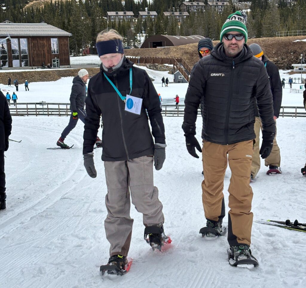 To personer iført vinterklær og truger går på en snødekt sti, med andre mennesker og bygninger i bakgrunnen.