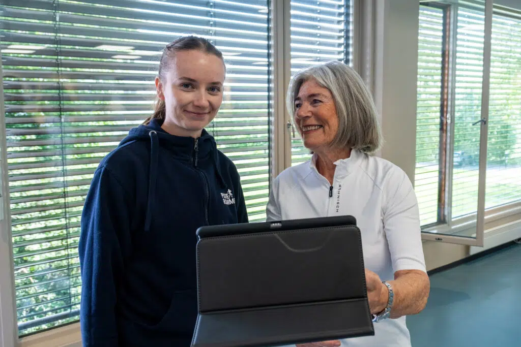 To kvinner står innendørs ved store vinduer; den ene holder et nettbrett og smiler, mens den andre vender seg mot kameraet med et nøytralt uttrykk.