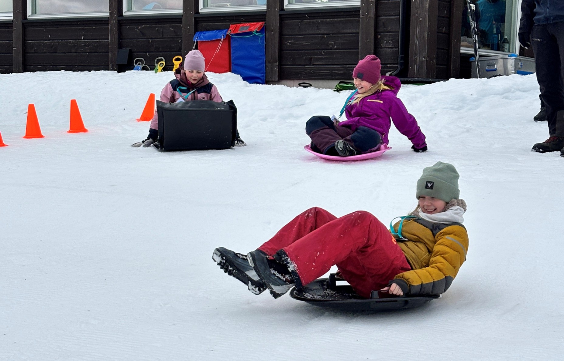 Tre barn aker ned en snødekt bakke i nærheten av oransje kjegler; to bruker runde kjelker, ett bruker en svart, rektangulær kjelke. En trebygning ligger i bakgrunnen.