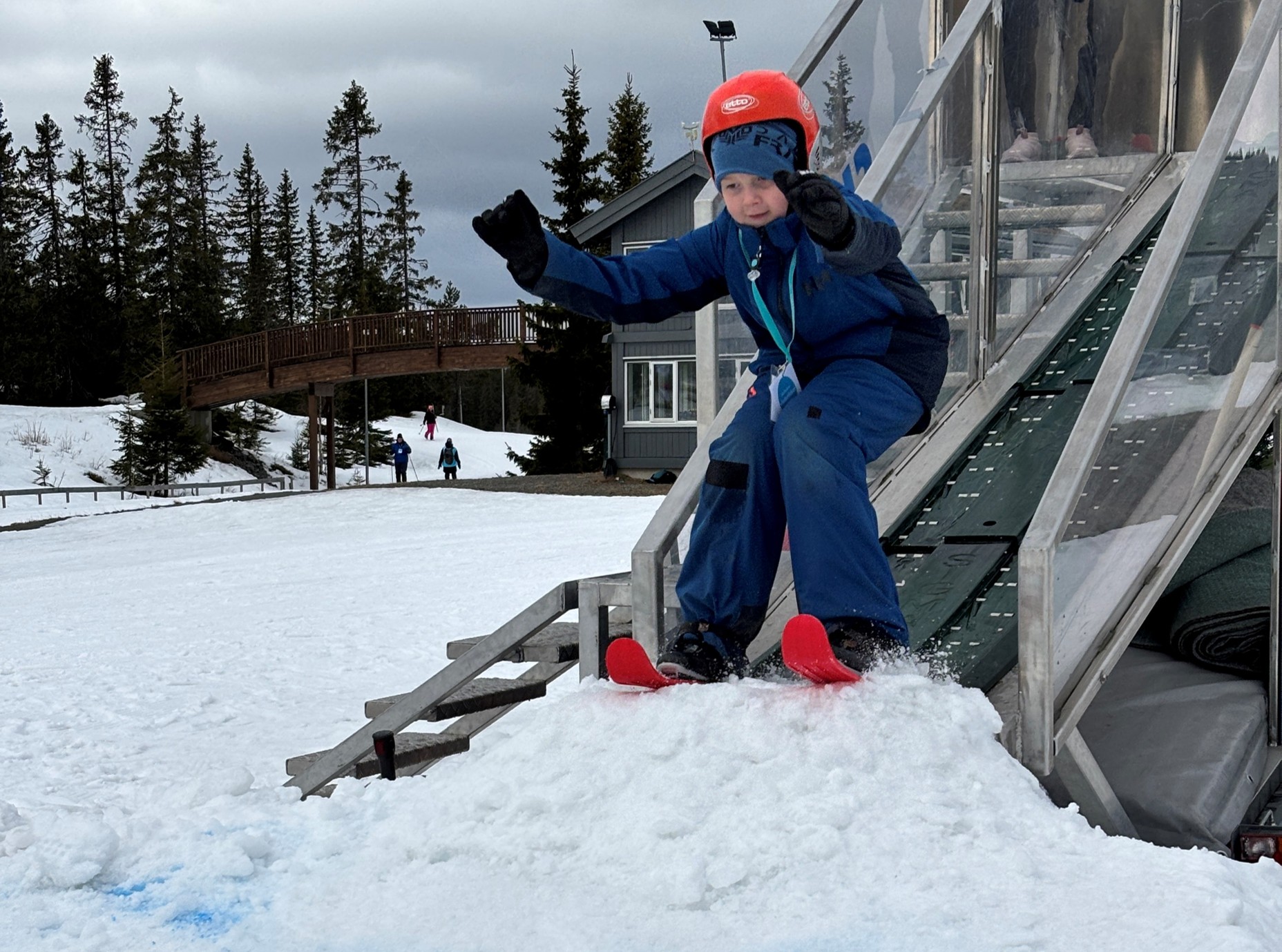 Et barn iført rød hjelm og blå snødress er i ferd med å stå på ski ned en snødekt rampe utendørs, med trær og en bygning i bakgrunnen.