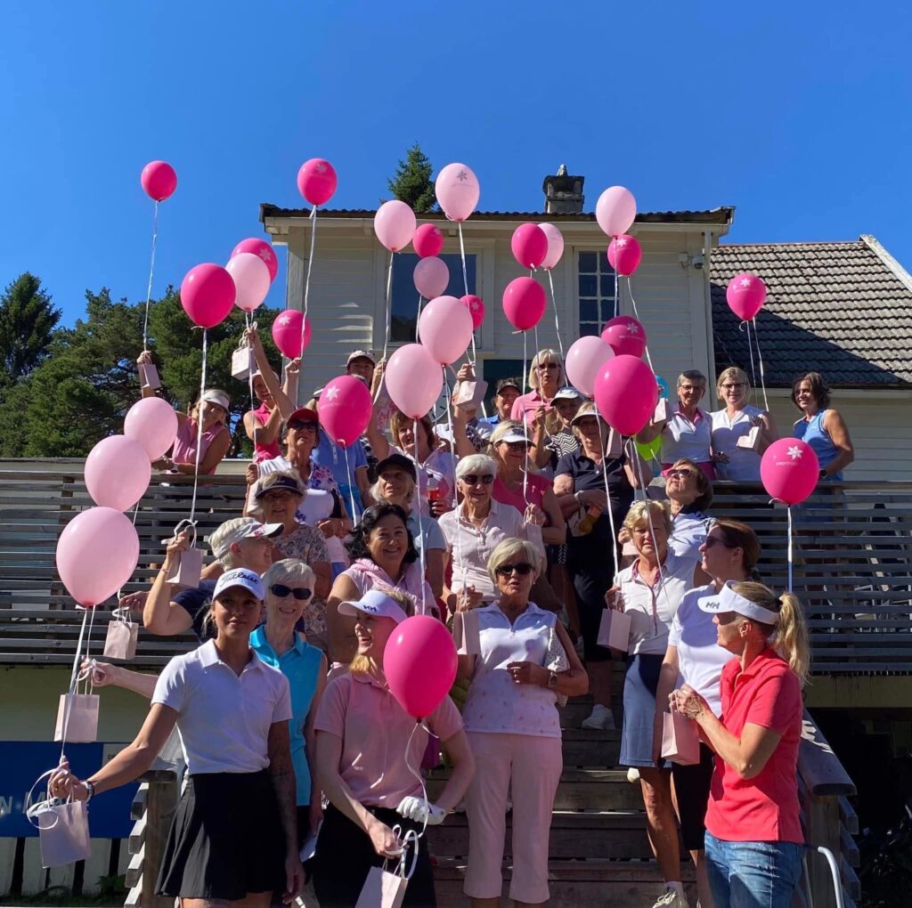 En gruppe kvinner står på en utendørs trapp med rosa ballonger og gaveposer i hendene og poserer for et gruppebilde foran en hvit bygning under en klarblå himmel, samlet for Et slag mot kreft.