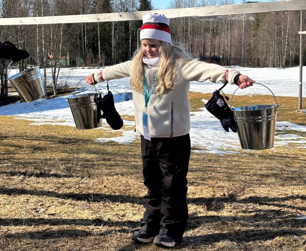 Et barn står utendørs med utstrakte armer og holder to metallbøtter, en i hver hånd, med snø og nakne trær i bakgrunnen.
