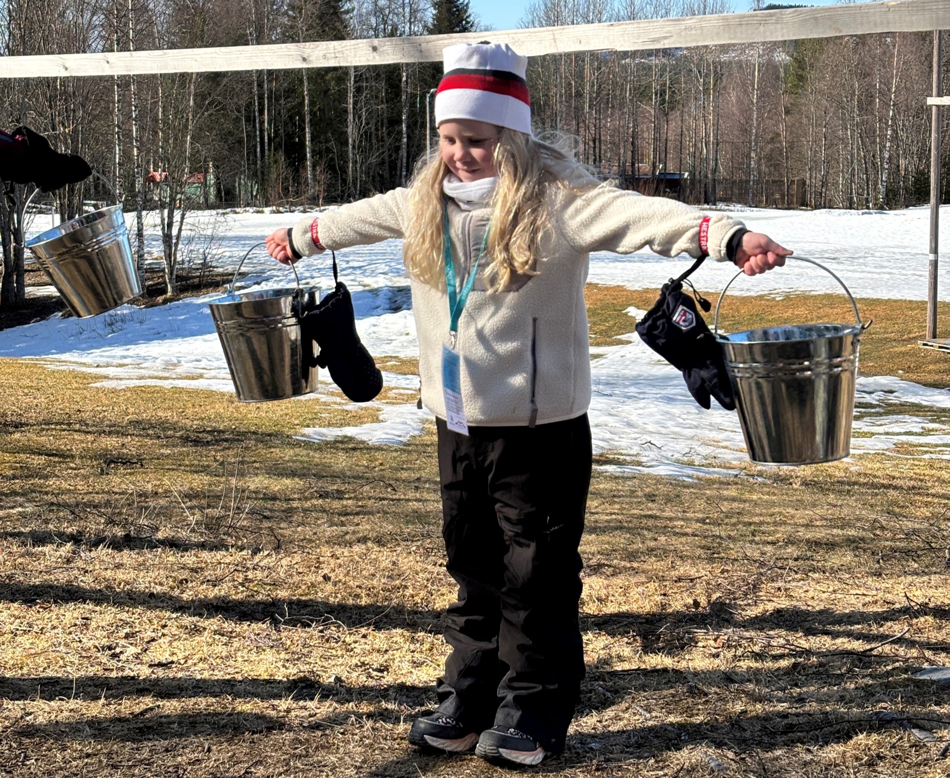 Et barn står utendørs med utstrakte armer og holder to metallbøtter, en i hver hånd, med snø og nakne trær i bakgrunnen.