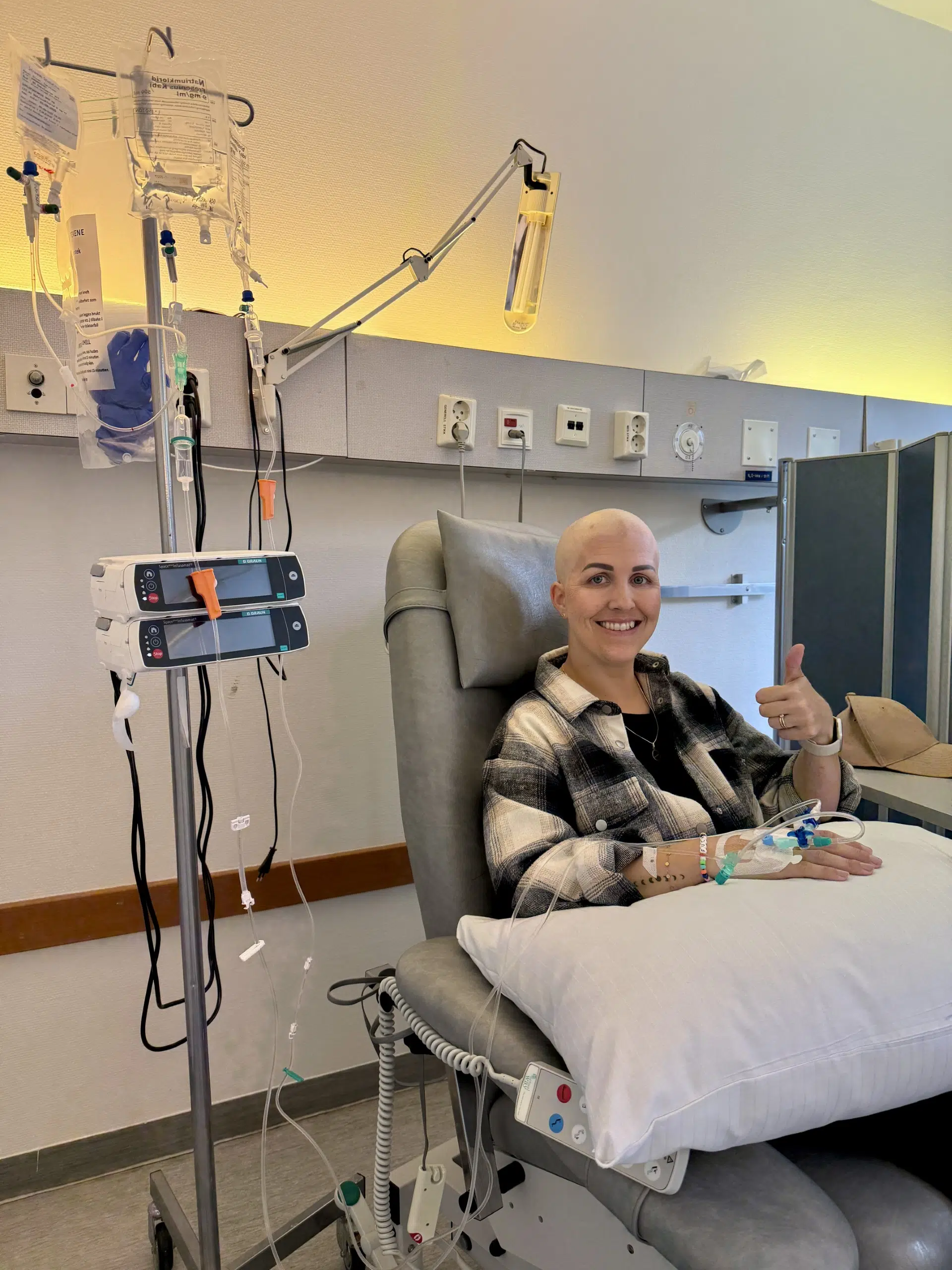 En person med barbert hode sitter i en sykehusstol og får intravenøs behandling, mens han smiler og gir tommelen opp. Medisinsk utstyr og infusjonsposer er synlige ved siden av dem.