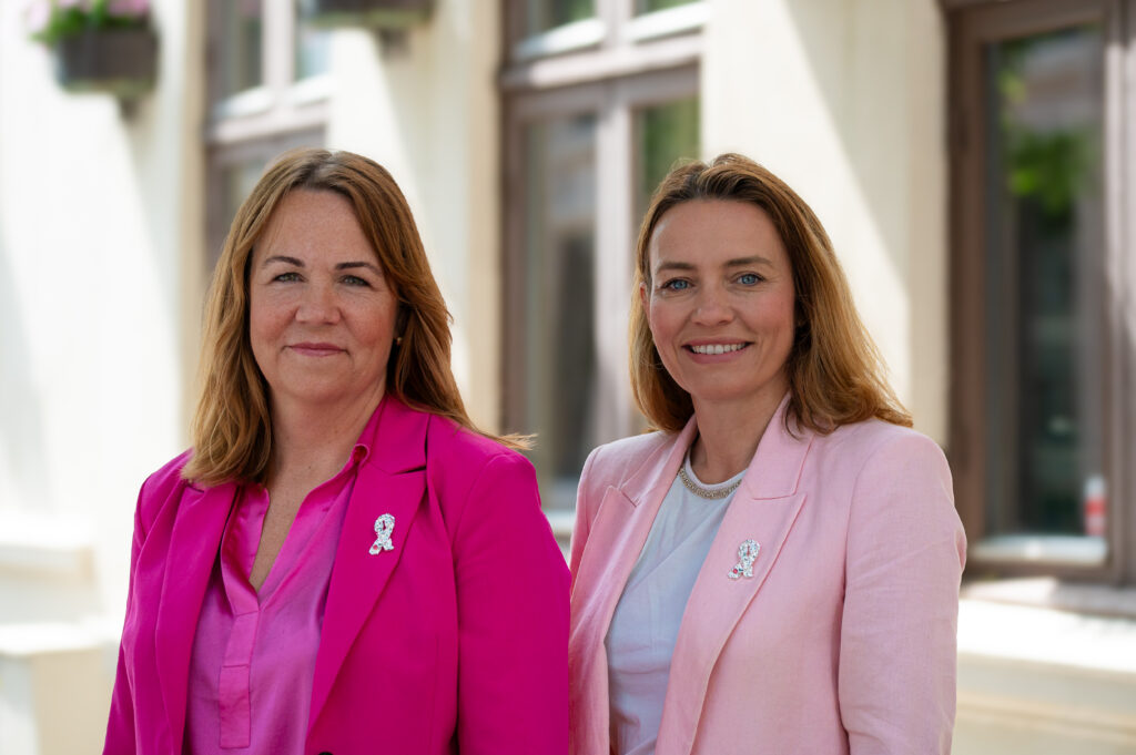 To kvinner iført rosa blazere og brystkreft-pins står side om side utendørs foran en bygning med store vinduer.