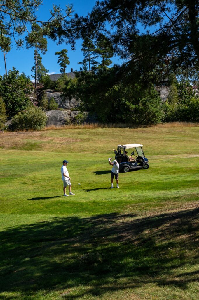 To personer står på en gressbelagt golfbane ved siden av en golfbil, med trær og en steinete skråning i bakgrunnen under en klarblå himmel.