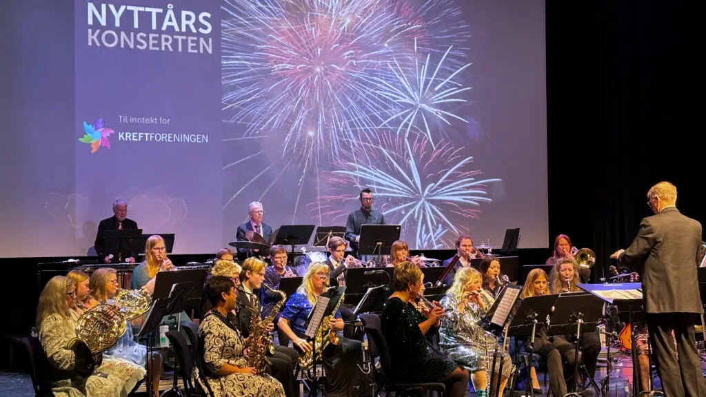 Et band spiller på scenen under et projisert bilde av fyrverkeri og teksten "Nyttårskonserten". En dirigent står foran de sittende musikerne.
