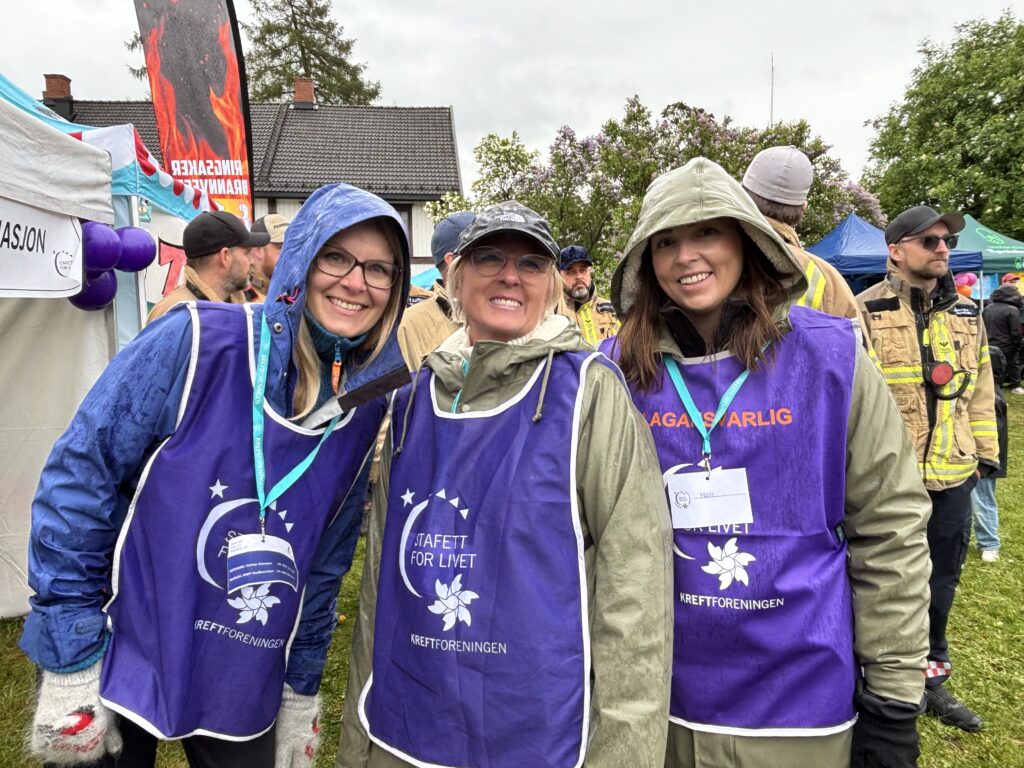 Tre kvinner iført lilla vester og regnjakker står utendørs på et arrangement og smiler til kameraet. Andre mennesker og telt er synlige i bakgrunnen.