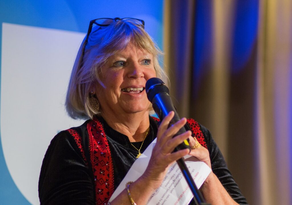 En kvinne med blondt hår og briller snakker i en mikrofon mens hun holder papirer, stående innendørs foran en blå og hvit bakgrunn.