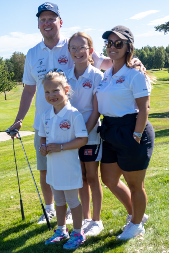 Fire personer i hvite poloskjorter står på en golfbane og smiler til kameraet; to voksne og to barn, hvorav den ene holder en golfkølle.