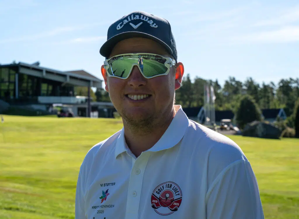 En mann med Callaway-cap, reflekterende solbriller og en hvit "Golf for Life"-polotrøye står på en golfbane med bygninger og trær i bakgrunnen.