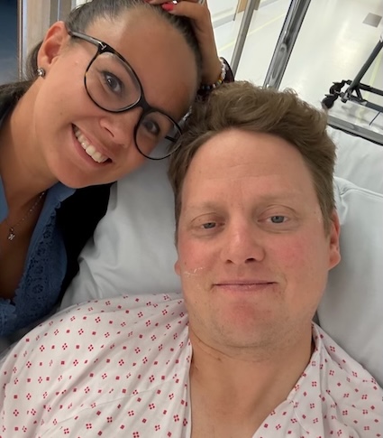 En kvinne smiler og poserer ved siden av en mann som ligger i en sykehusseng, iført sykehuskjole.