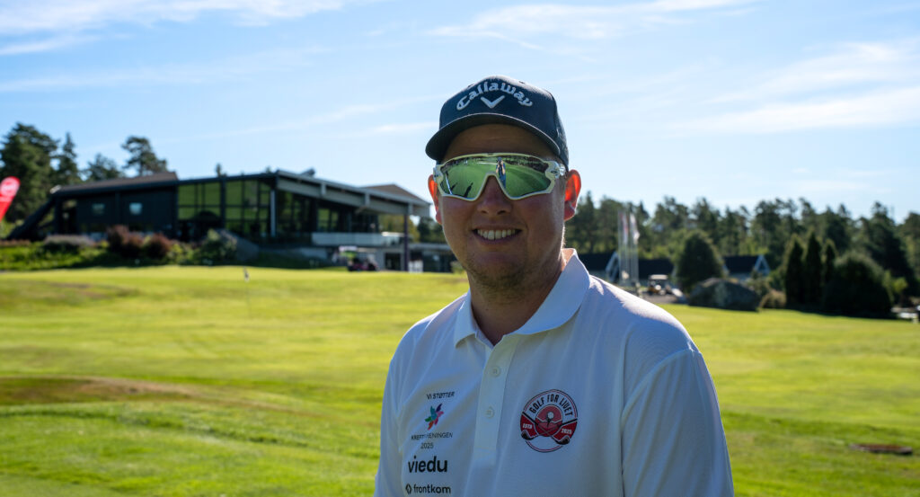 En mann med solbriller, Callaway-lue og hvit poloskjorte står på en golfbane med et klubbhus og trær i bakgrunnen.