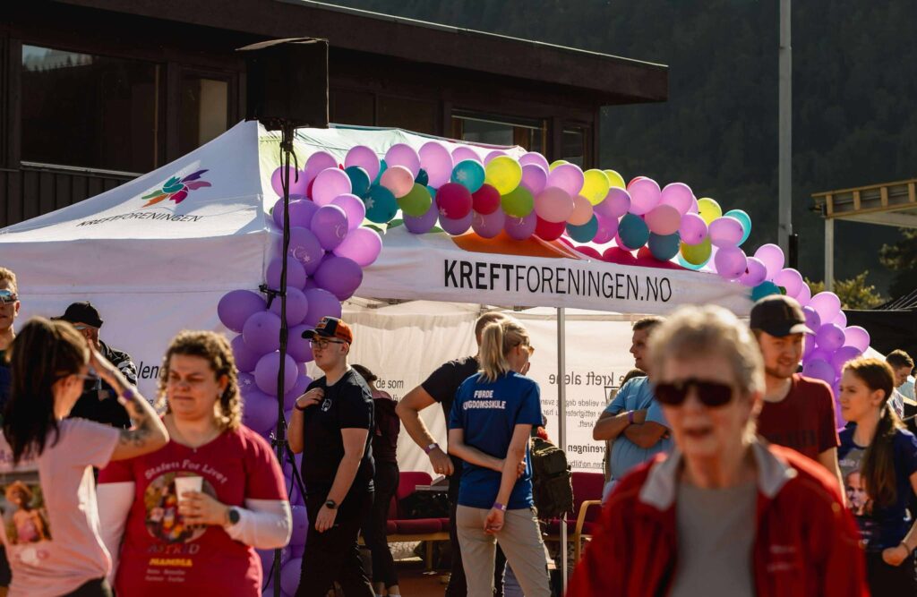 En folkemengde samler seg ved et hvitt telt dekorert med fargerike ballonger og merket med "Kreftforeningen.no" på et utendørsarrangement.