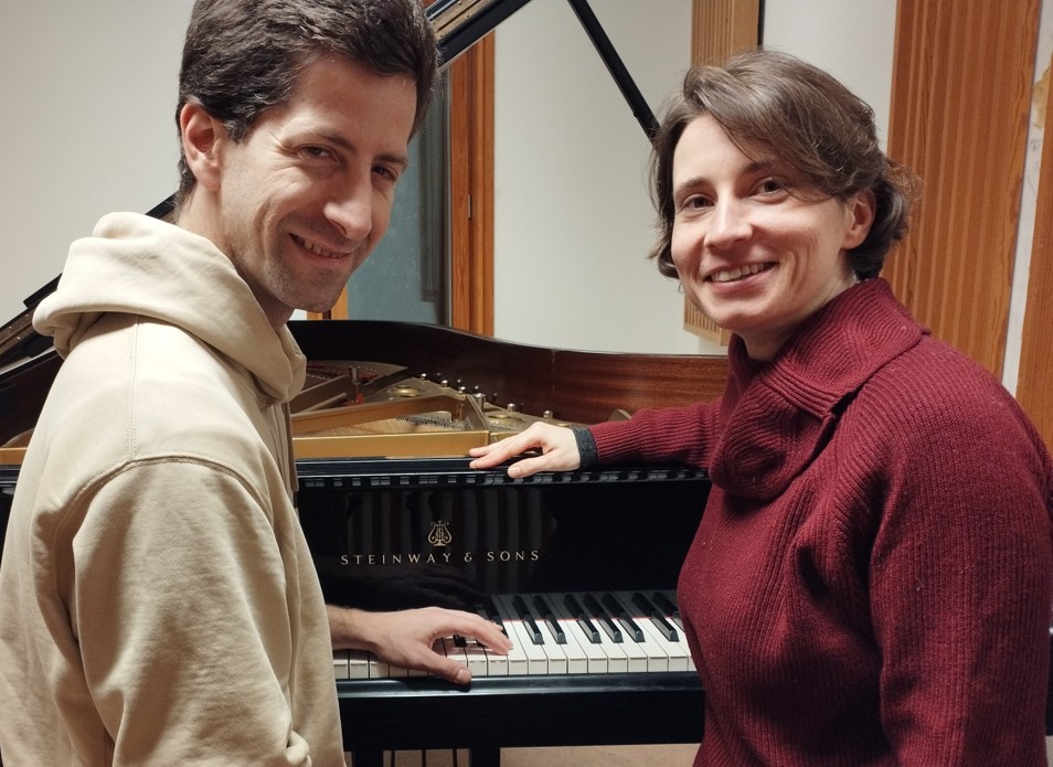 To personer står smilende ved siden av et Steinway & Sons-flygel, med den enes hånd hvilende på tangentene.