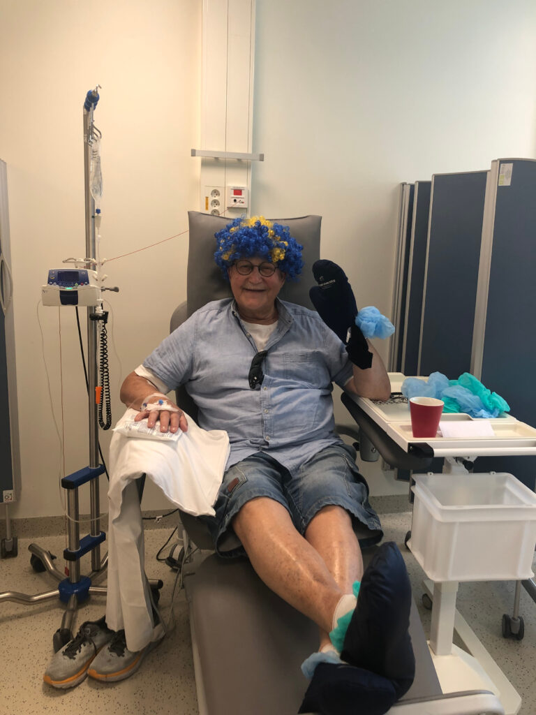 En mann i en sykeseng får intravenøs behandling, iført blå og gul parykk, svart grytevott og fritidsklær i et sykehusmiljø.