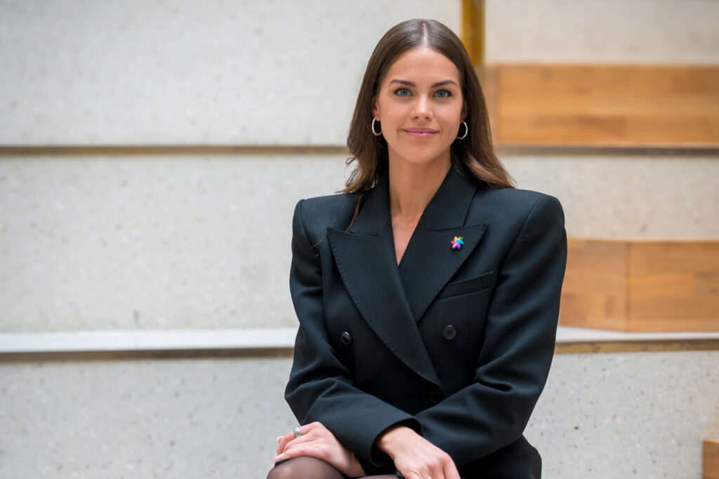 En kvinne med langt brunt hår, iført svart blazer og øreringer, sitter på en innendørstrapp med hendene foldet på kneet.