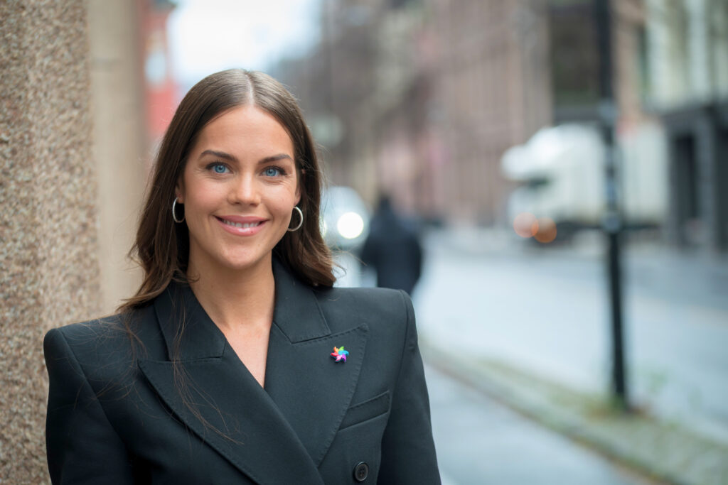 En kvinne med langt, brunt hår, iført svart blazer og stjernenål, står og smiler utendørs i en bygate.