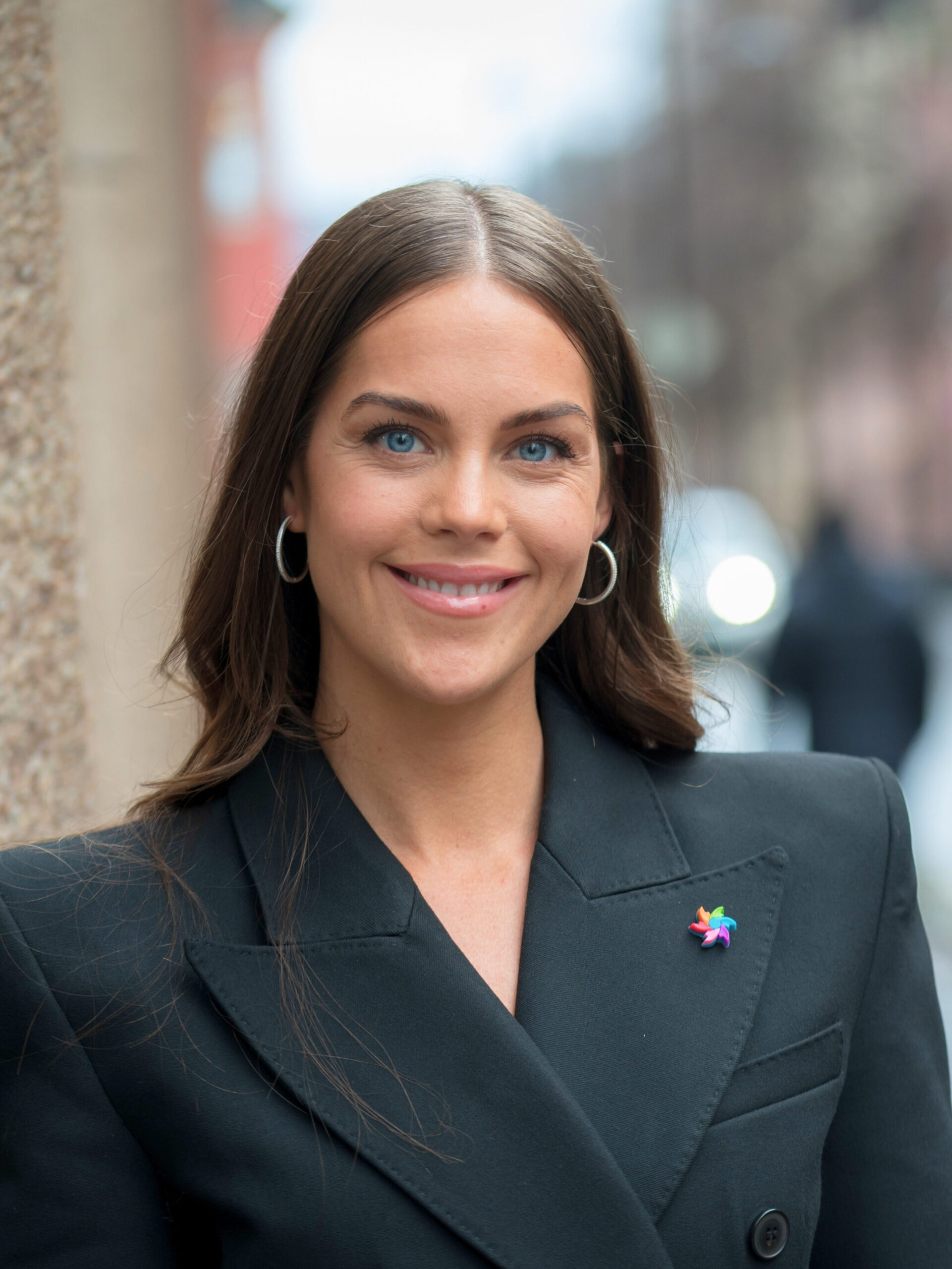 En kvinne med langt, brunt hår, iført svart blazer og stjernenål, står og smiler utendørs i en bygate.