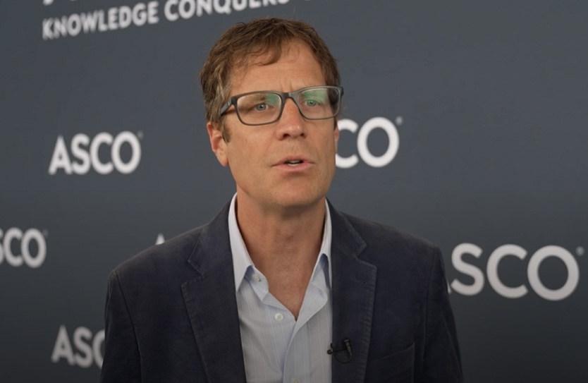 En mann med briller og blazer taler foran et bakteppe med ASCO-logoen og frasen "Knowledge Conquers".