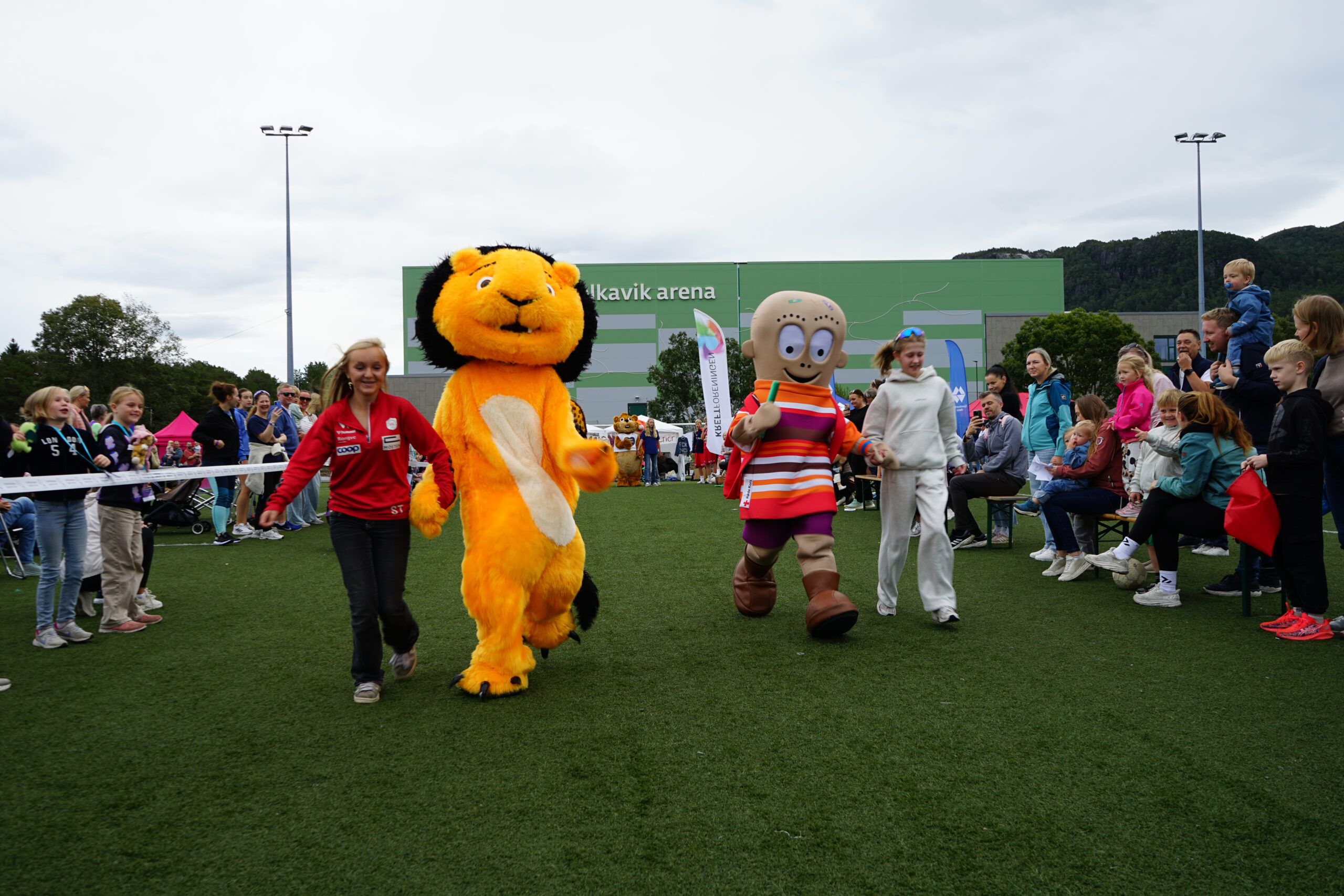 To personer i tegneseriefigurkostymer går sammen med to voksne på en idrettsplass, omgitt av en folkemengde av barn og voksne på et utendørsarrangement.