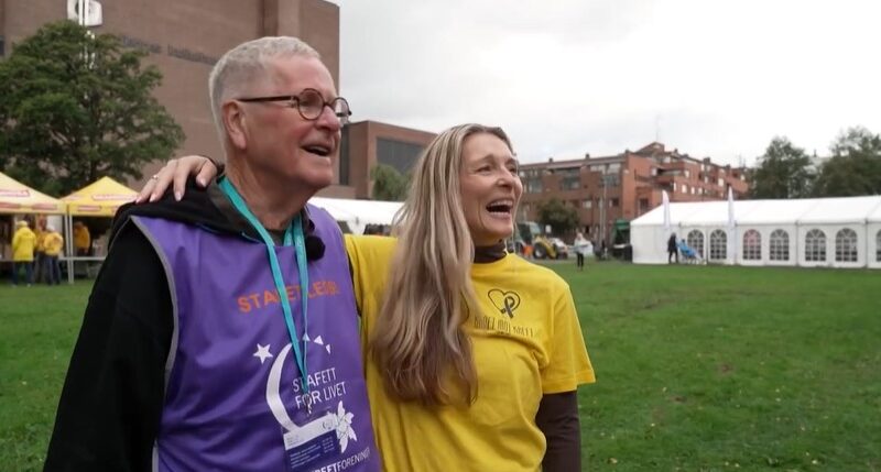 To personer iført event-skjorter står smilende sammen på en gresslette, med telt og bygninger i bakgrunnen.