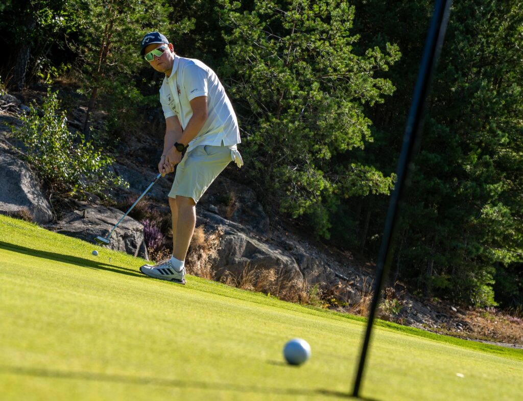 En person i solbriller og hvitt antrekk putter en golfball på en green, med trær og steiner i bakgrunnen.
