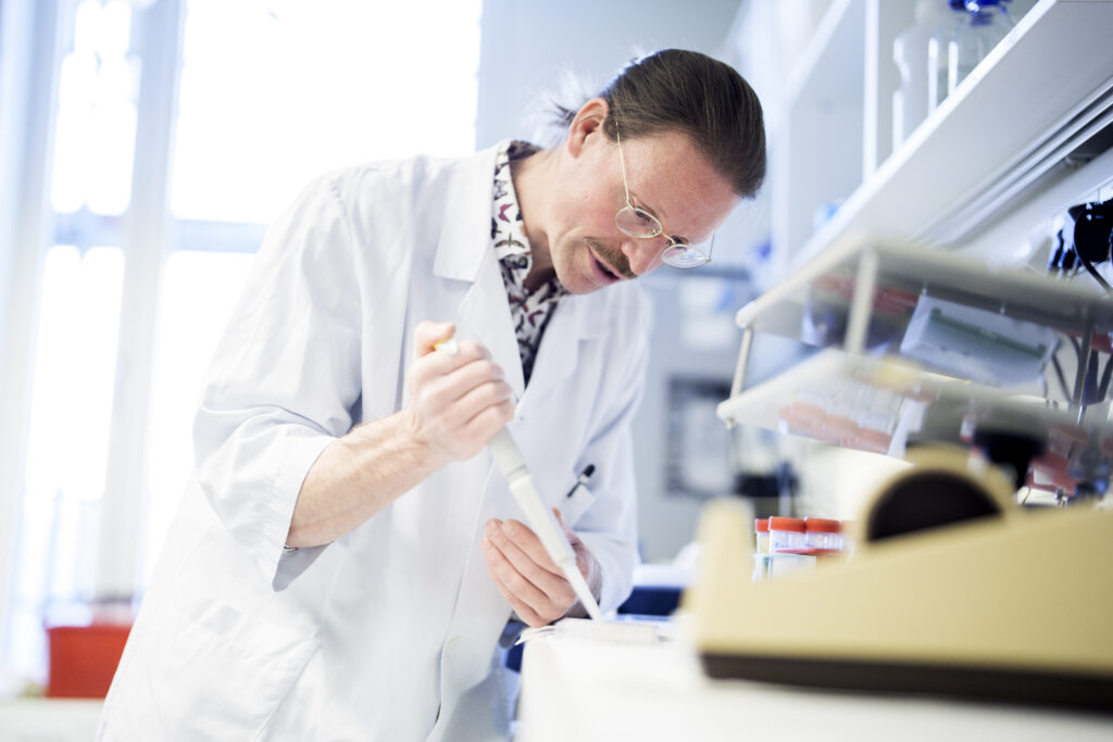 En forsker i hvit laboratoriefrakk bruker en pipette til å overføre væske i et laboratorium med utstyr og rekvisita på benken.