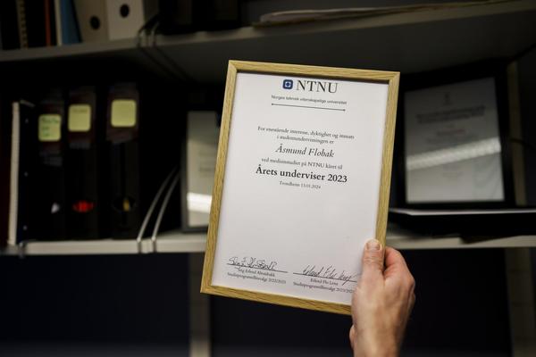 En hånd holder et innrammet diplom fra NTNU som Åsmund Flobak har fått for å være "Årets underviser 2023", med hyller og mapper i bakgrunnen.