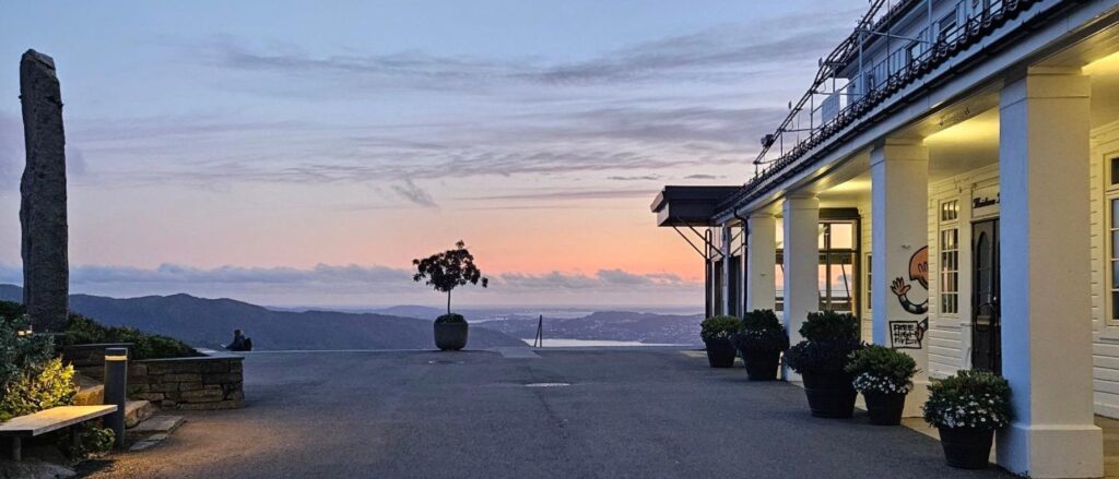 En bygning med store søyler og potteplanter står i nærheten av et naturskjønt utsiktspunkt i solnedgangen, med åser, vann og en pastellfarget himmel i bakgrunnen.