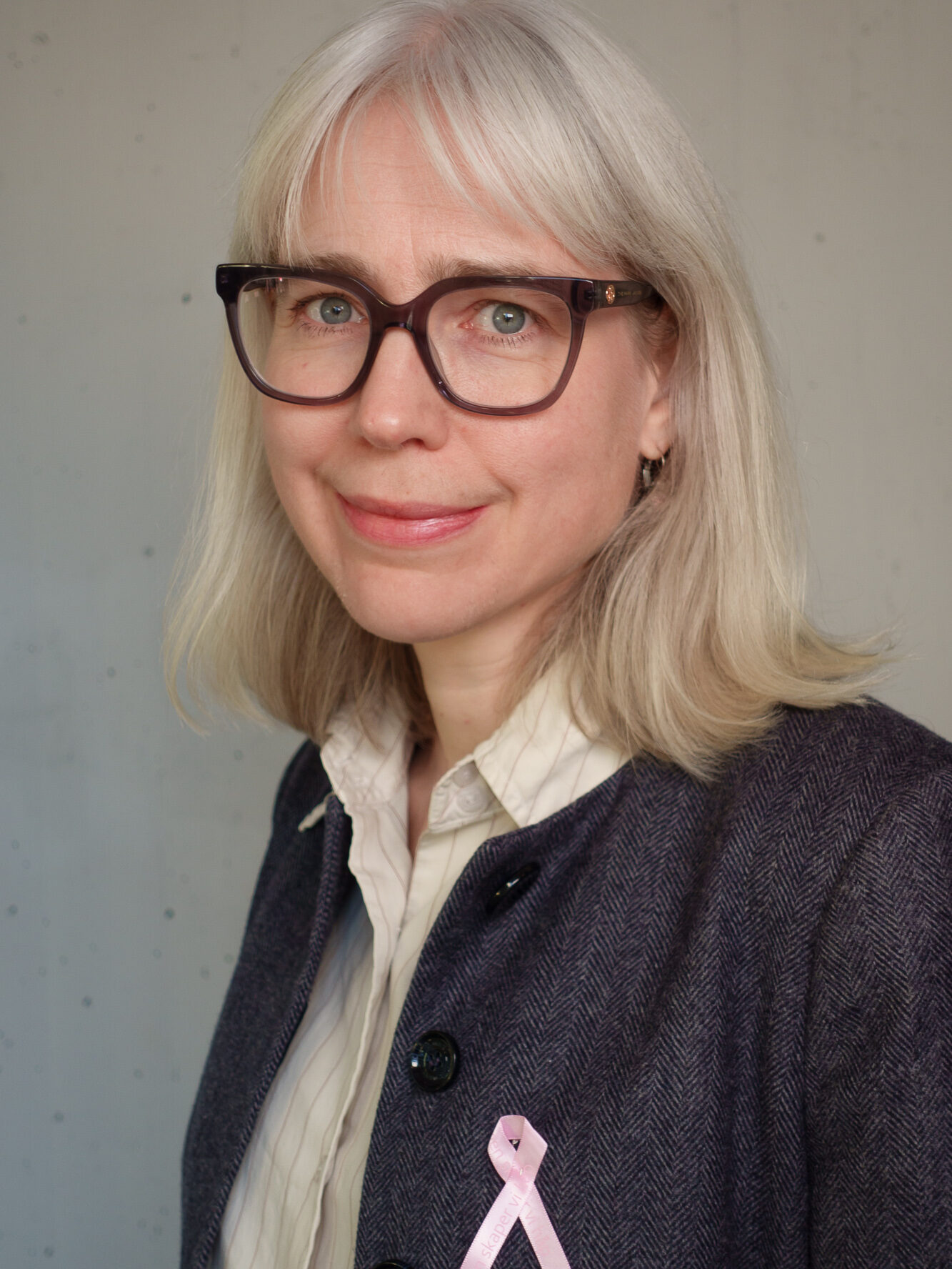 En kvinne med grått hår og briller bærer en dressjakke med et rosa bånd festet på, og står mot en ensfarget, lys bakgrunn.