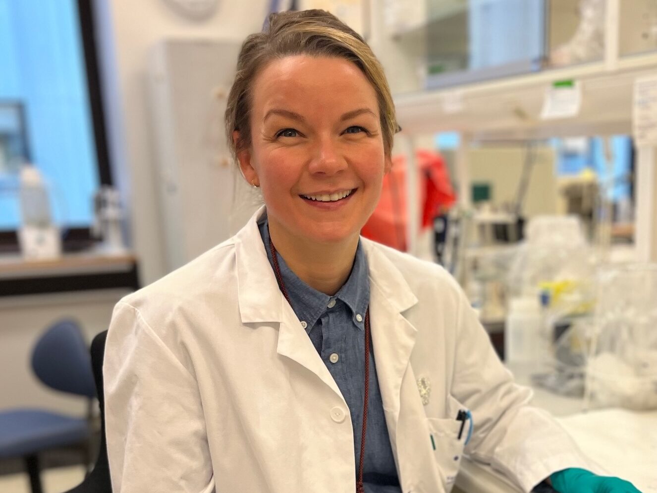 En person i hvit laboratoriefrakk, blå skjorte og blågrønn hanske sitter og smiler i et laboratorium med vitenskapelig utstyr i bakgrunnen.