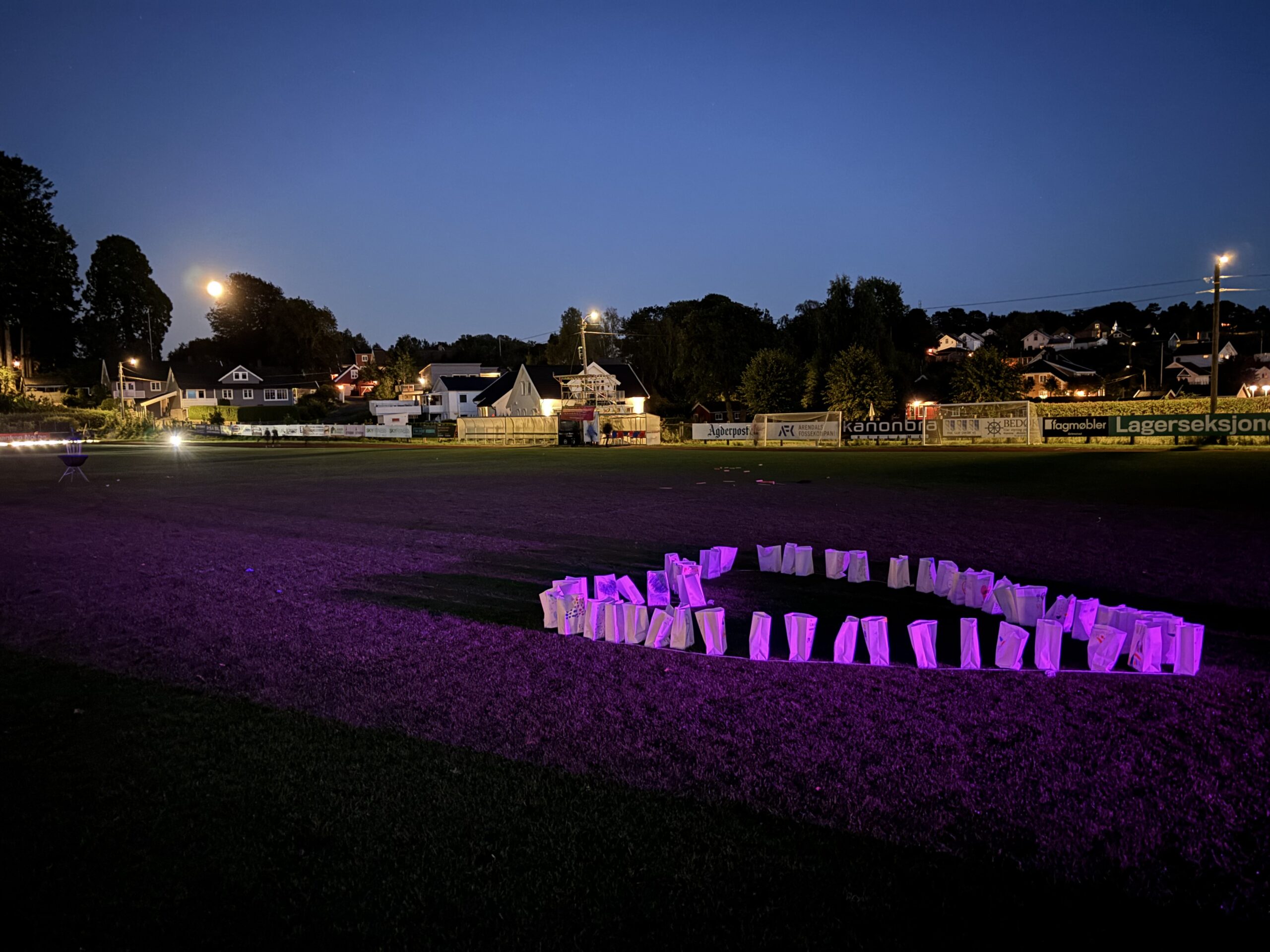 En idrettsplass i skumringen med et sirkelformet arrangement av lysende hvite papirposer som lyser lilla i forgrunnen. Hus og trær er synlige i bakgrunnen.