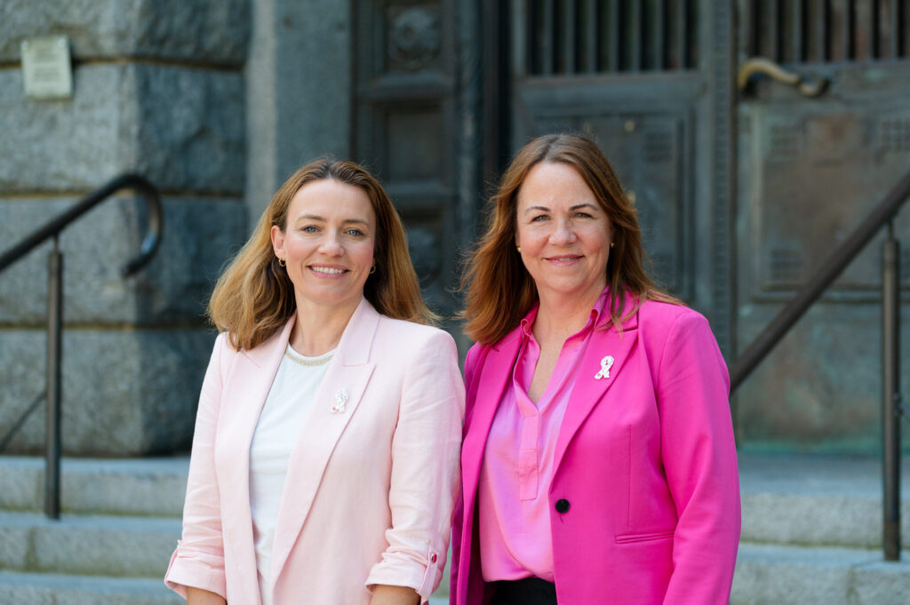 To kvinner står utenfor en bygning, begge iført rosa blazere og brystkreftsløyfer, vendt mot kamera og smilende.
