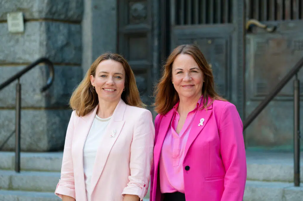 To kvinner står utenfor en bygning, begge iført rosa blazere og brystkreftsløyfer, vendt mot kamera og smilende.